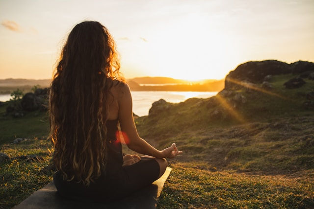 Woman with long hair meditates on a grassy hill at sunset, facing a glowing lake and rocky shoreline.