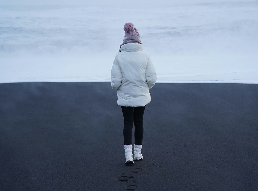 Person in white coat and pink hat walks on black sand beach. Ocean waves in the background create a calm, serene mood.