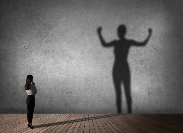 A woman stands on wooden floor, looking at her shadow flexing muscles on a gray wall, suggesting inner strength and empowerment.