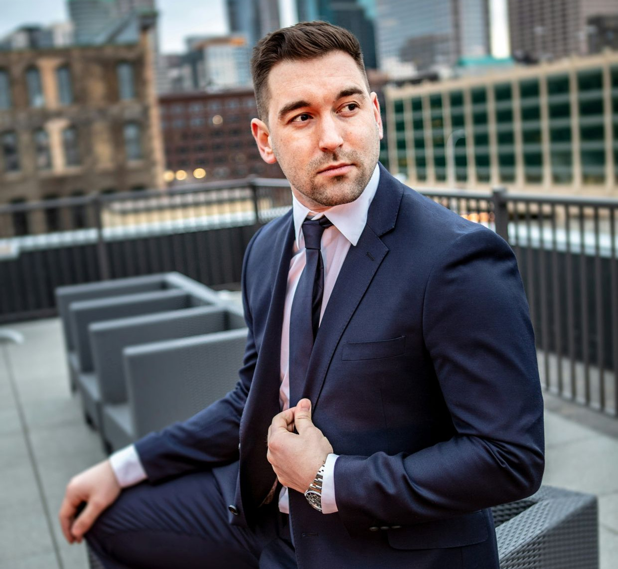 Suited man adjusts his tie on a rooftop terrace, with a blurred city skyline behind him and a serious expression.
