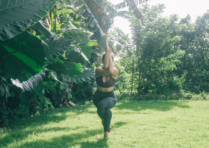 A woman in workout gear practices yoga on a sunny lawn, surrounded by lush green foliage. The scene is peaceful and natural.