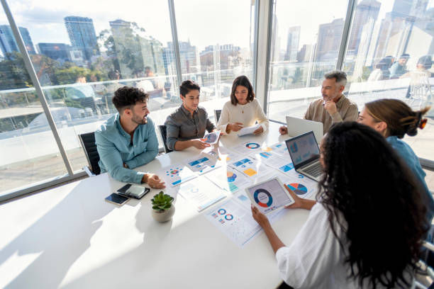 People sit around a bright office table with charts and laptops, discussing and reviewing documents. Cityscape visible through windows.