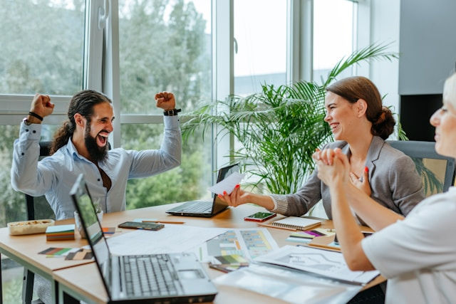A team of three colleagues celebrates a successful project, with one person raising their arms in triumph.