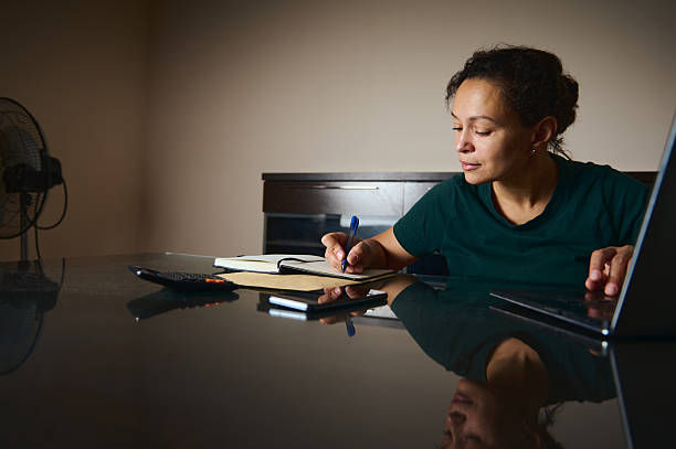 Woman in a green shirt writes in a notebook at a glossy table, with a laptop and fan nearby in a dim room.