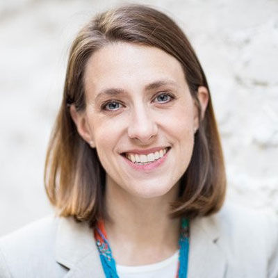 Smiling woman with shoulder-length brown hair, wearing a light blazer and colorful scarf. Neutral, textured background.
