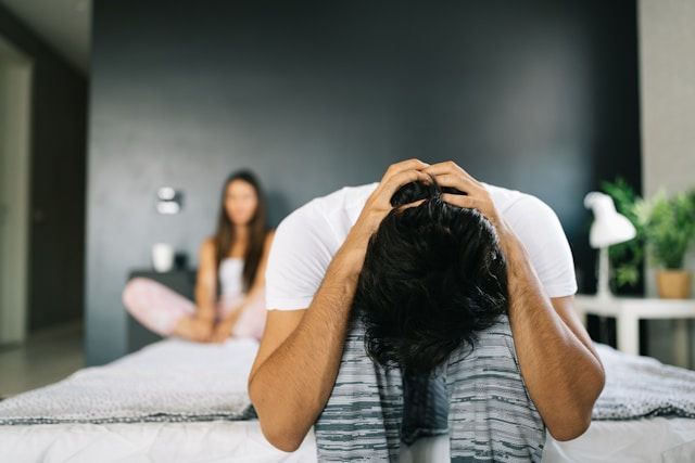Distressed man sits on bed with head in hands, while a blurred woman sits behind him in a modern bedroom.