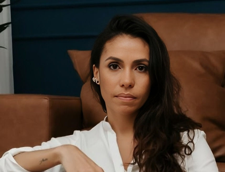 Woman with long dark hair in a white blouse sits on a brown leather couch, looking calmly at the camera.