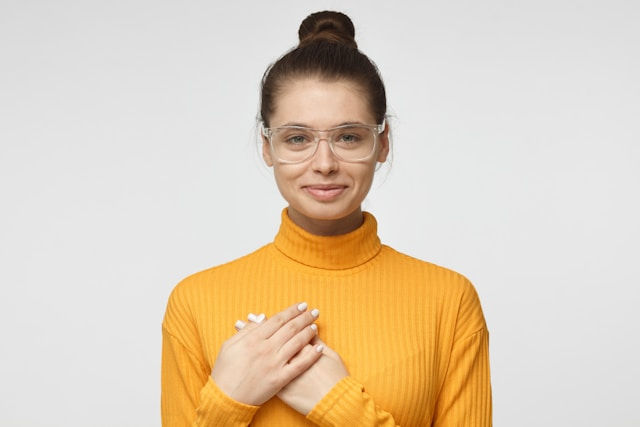Woman in orange turtleneck and glasses, smiling with hands on chest against plain background. Calm mood, neutral setting.