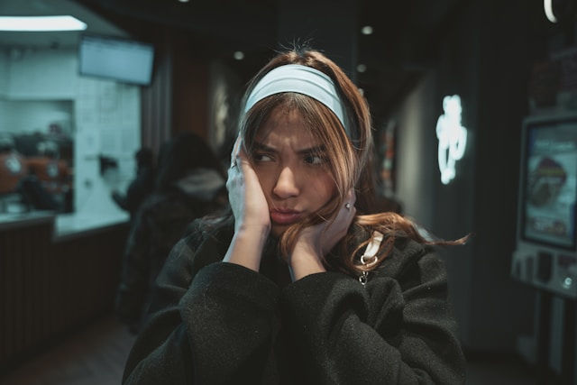 A young woman stands indoors with a frustrated expression, pressing her hands against her cheeks while wearing a light blue headband.