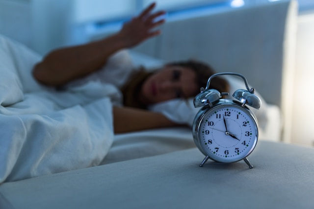 Silver alarm clock showing 4:00 AM on bedside table with a woman lying in bed, reaching towards it, in a softly lit bedroom.