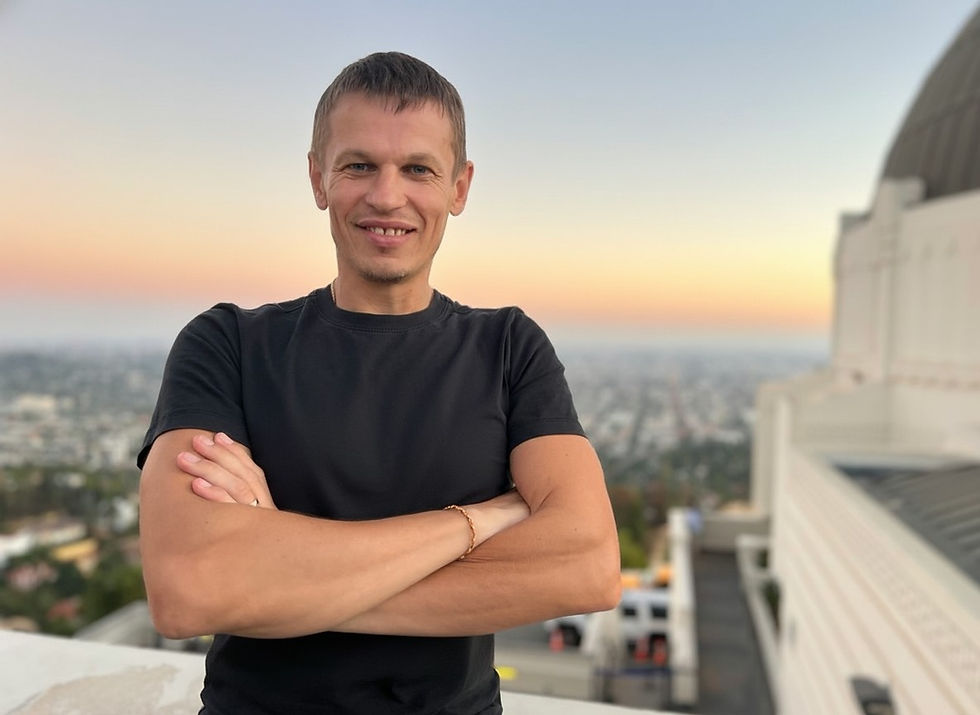 Smiling person in a black shirt stands with crossed arms at sunset, city view and domed building in the blurred background.