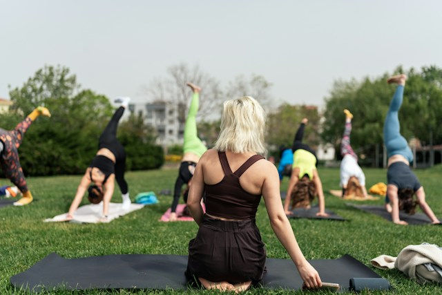 Outdoor yoga class in a park, with a blonde woman seated on a mat watching others do handstands on a green lawn.