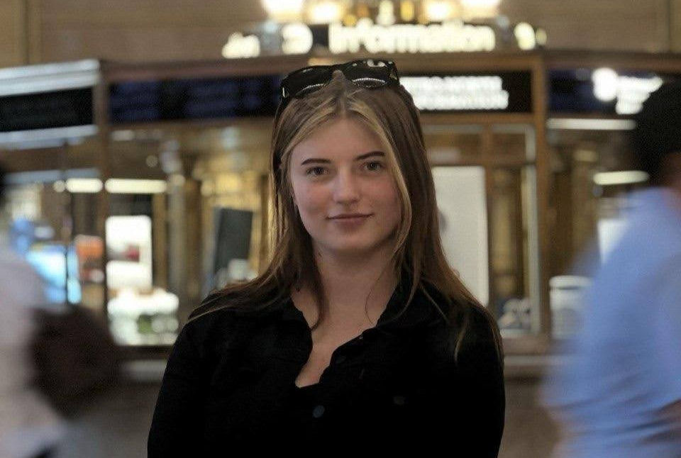 Young woman in black shirt with sunglasses on head smiles in a busy indoor mall, with blurred signs and people behind her.