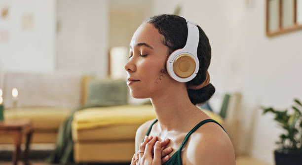 Woman in white headphones meditates with eyes closed and hands on chest in a cozy living room.