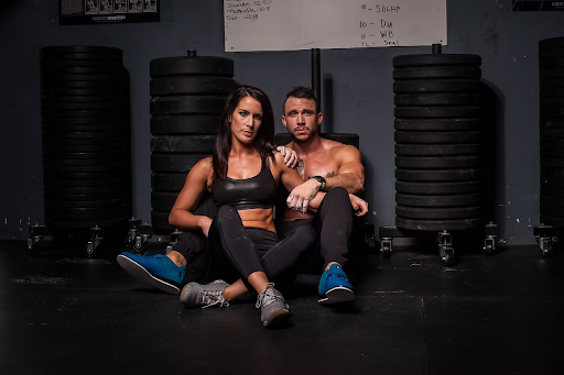 A fit couple in black workout gear sits on a gym floor with stacked weight plates behind them. They appear relaxed and confident.
