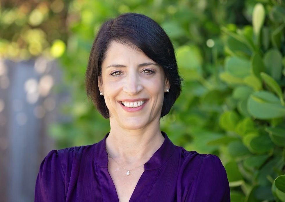 Smiling woman with short dark hair in a purple blouse, stands in front of lush green foliage, exuding a warm, confident mood.