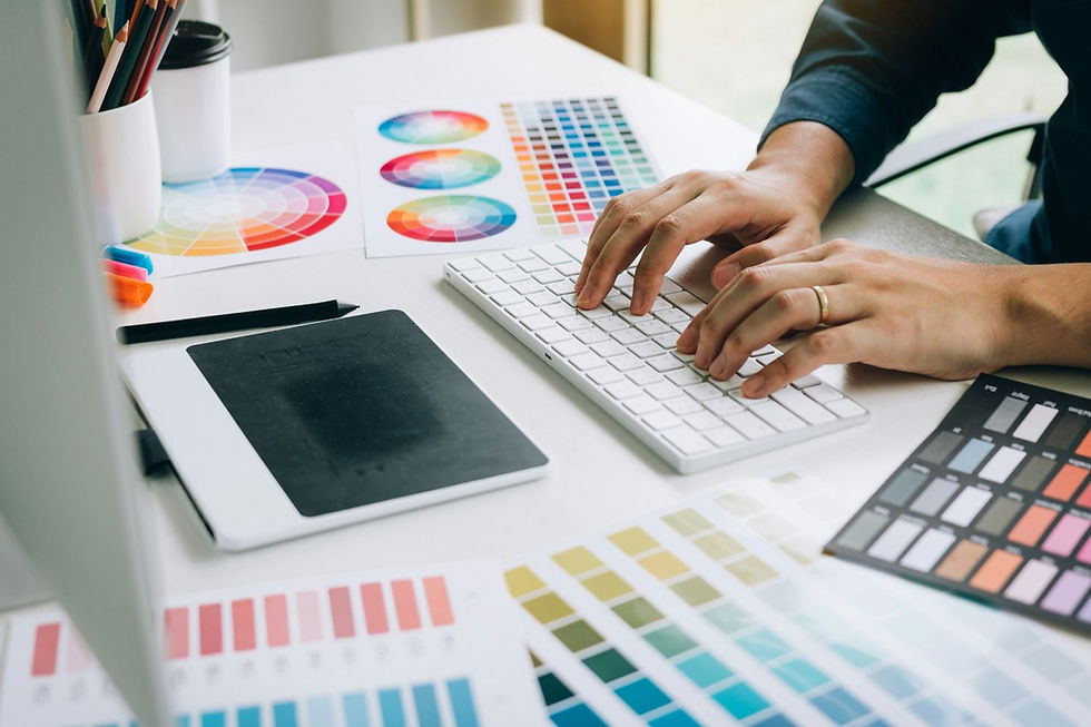 Hands typing on a keyboard surrounded by color swatches, a tablet, pens, and a coffee cup on a white desk, suggesting creativity.