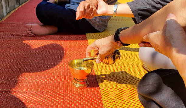 Hands pour water from a cup onto a mat in a ritual ceremony. The setting includes a red and yellow woven mat, with sunlight casting shadows.
