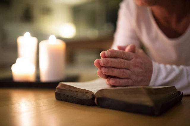 A person is praying with their hands clasped over an open book, with candles lit in the background.