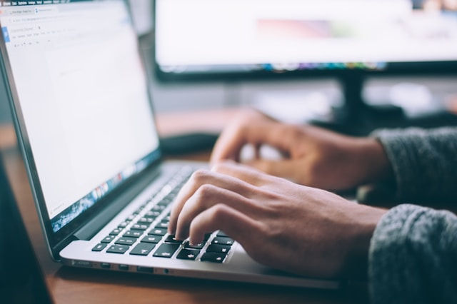 The photo shows a person typing on a laptop, with a blurred background of computer screens displaying documents and other content.