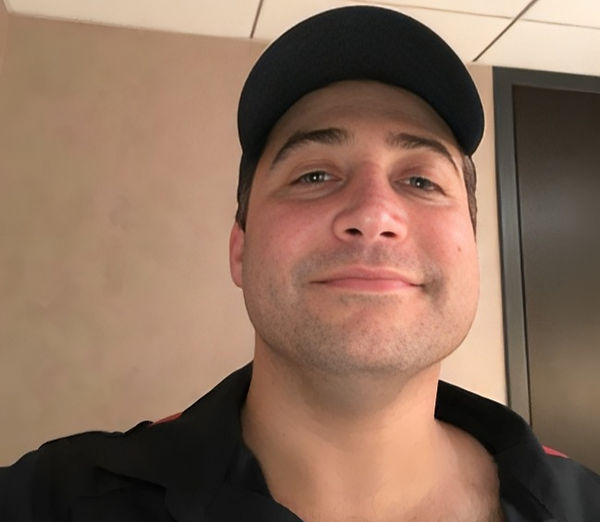 Man wearing a navy cap smiles slightly in a beige room. The background features a ceiling and a dark door. The mood is neutral.