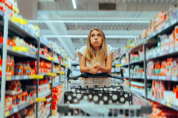 A young woman stands with a shopping cart in a grocery aisle, looking up with a puzzled or thoughtful expression.