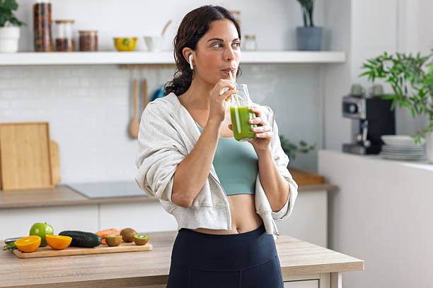 Woman in a bright kitchen sips a green smoothie from a glass jar, with fruit on the counter and a relaxed, healthy mood.