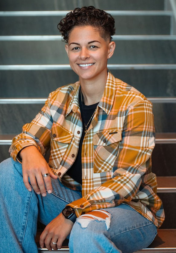 Person with short curly hair sits on stairs, smiling. Wearing a yellow plaid shirt and jeans. Relaxed mood, neutral background.