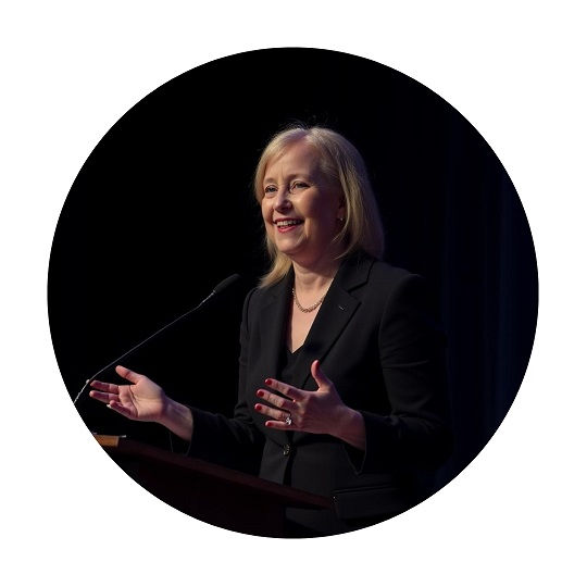 A woman in a black suit speaks passionately at a podium against a dark background, gesturing with her hands and smiling warmly.