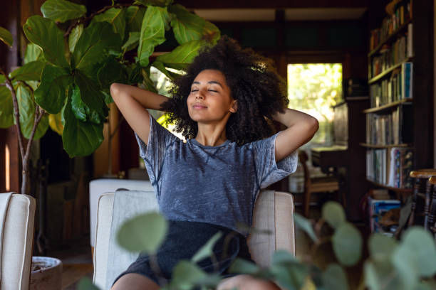 A woman sits peacefully in a cozy room with her hands behind her head and eyes closed, enjoying a moment of relaxation surrounded by greenery and books.