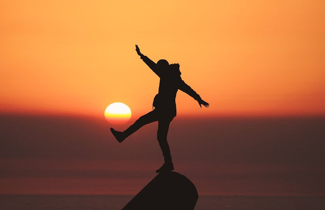 Silhouette of a person balancing on one leg atop a rock at sunset, arms outstretched against an orange sky and sun.