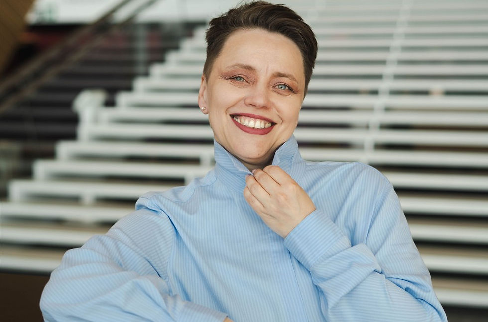 Smiling person in a light blue striped shirt adjusts their collar in front of a blurred staircase.