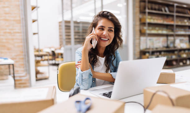 Smiling woman on phone at a laptop in a bright office warehouse, with cardboard boxes around her.