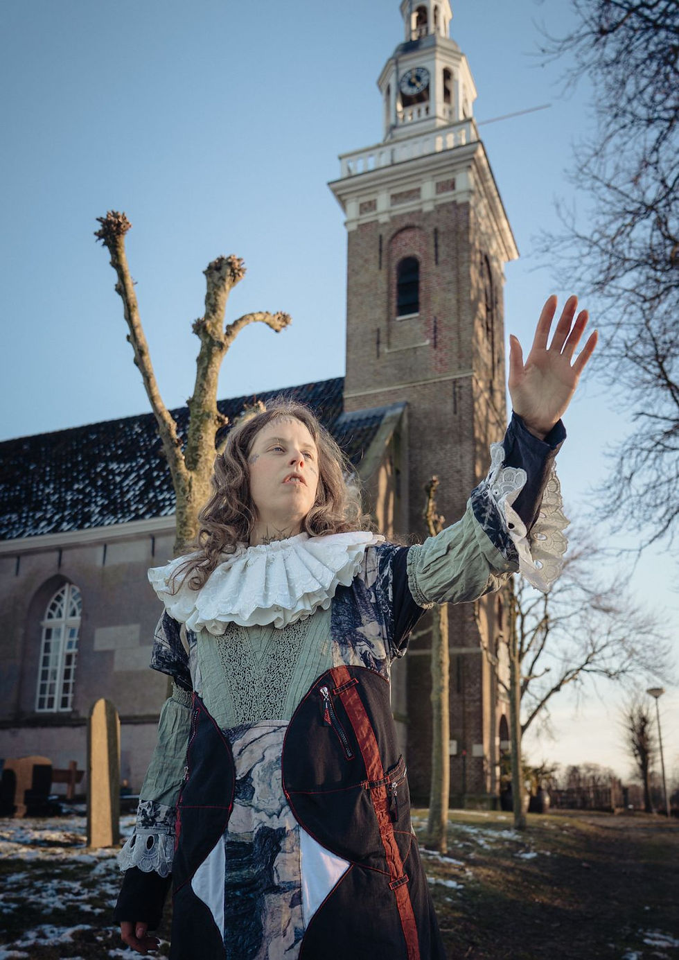 Person in ornate lace attire, arms reaching forward, with a tall brick tower background and bare branches against a clear sky. Moody atmosphere.