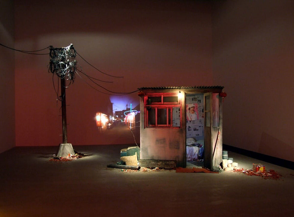 A dimly lit room shows a small, rundown shack with a red window, lit bulb, and messy interior. Nearby, a tangled wire pole. A street scene is projected.