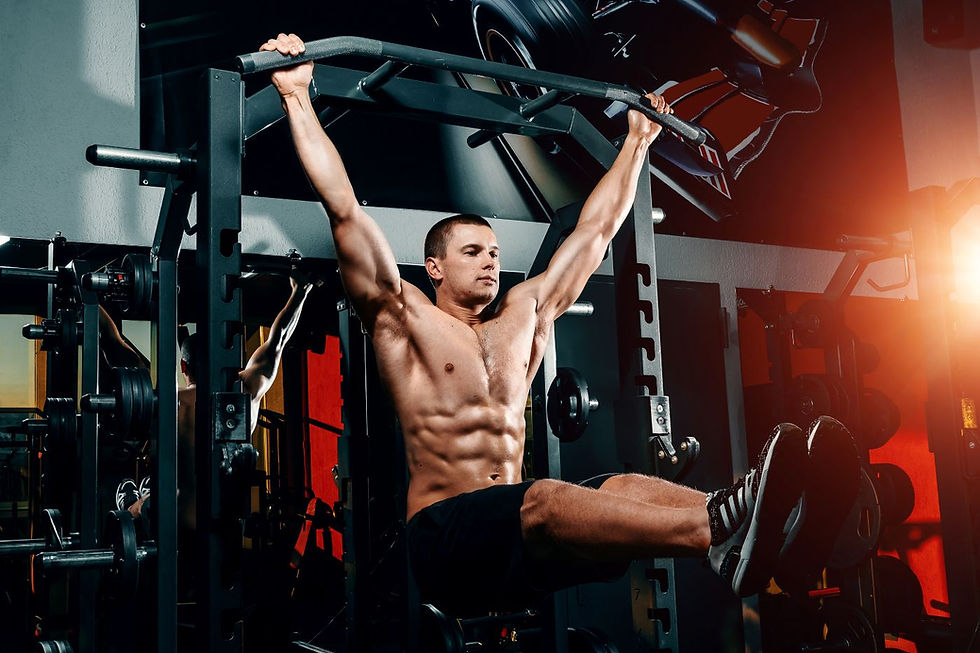 Muscular man doing leg raises on a pull-up bar in a gym. He is shirtless, wearing black shorts, with gym equipment and red-orange lighting.