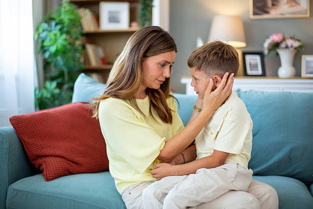 Mother gently comforts a young boy on a blue couch in a cozy living room, both in pale yellow, warm and caring.