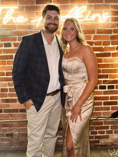 Couple smiling in front of a brick wall with neon sign. Man in plaid jacket, woman in satin dress. Warm lighting, relaxed atmosphere.