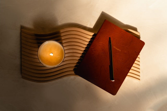 Candle and journal with a pen on a wavy wooden surface. Warm lighting creates a cozy mood. Journal features abstract face design.