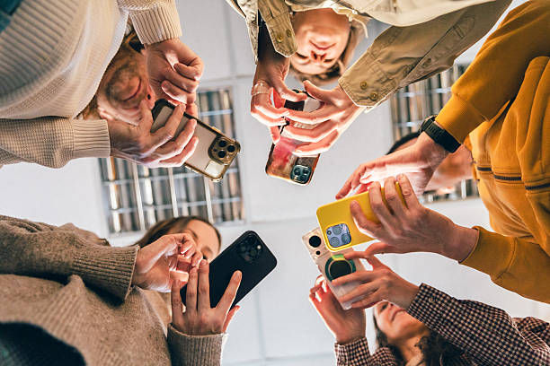 Group of friends huddled in a circle, looking down at their smartphones indoors, with bright clothing and a lively mood.