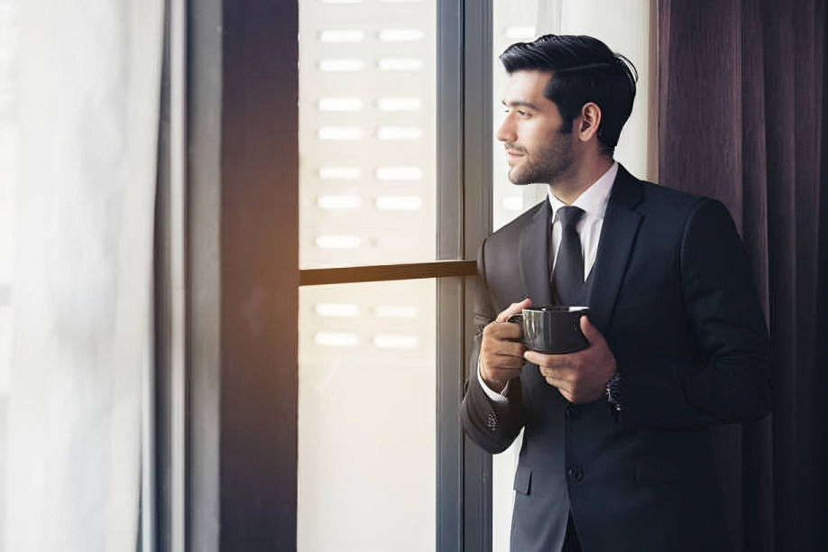 Man in a black suit holds a mug, gazing out a window. Sunlight enters, creating a serene mood. Room has white curtains and dark walls.