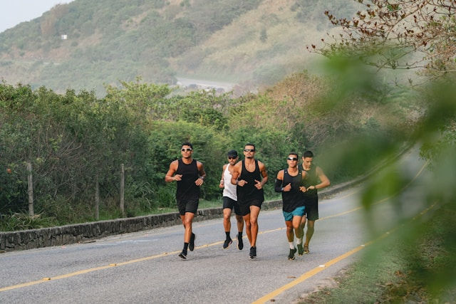 Five people in athletic gear jogging on a country road; lush greenery surrounds them. They appear focused and in-sync on a clear day.