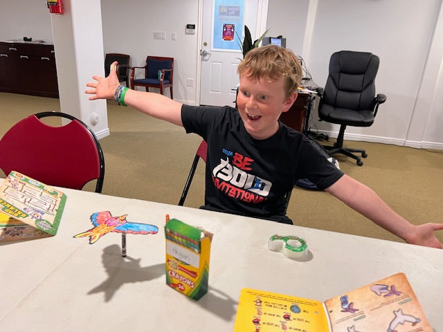 Excited boy in a black T-shirt sits at a table with crafts and crayons in an office room, arms spread wide and smiling.