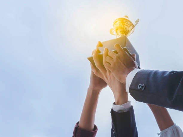 Hands lift a trophy toward a glowing idea icon against a bright blue sky, symbolizing success and inspiration.