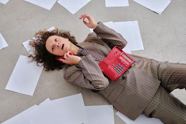 Person in brown pinstripe suit lies on office floor among scattered papers, talking on a red phone with a stressed look.