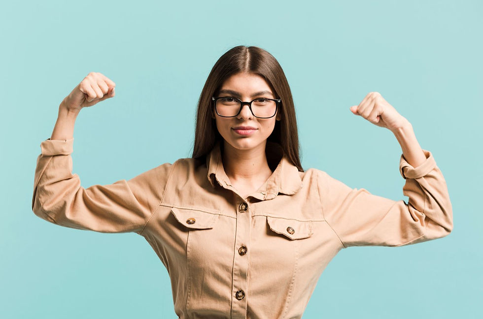 A confident young woman wearing glasses and a beige shirt is flexing her arms in a strong pose against a light blue background.