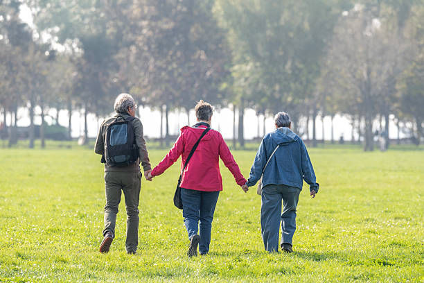 Three people walk hand in hand across a green park, backs turned, with trees and soft morning light in the background.