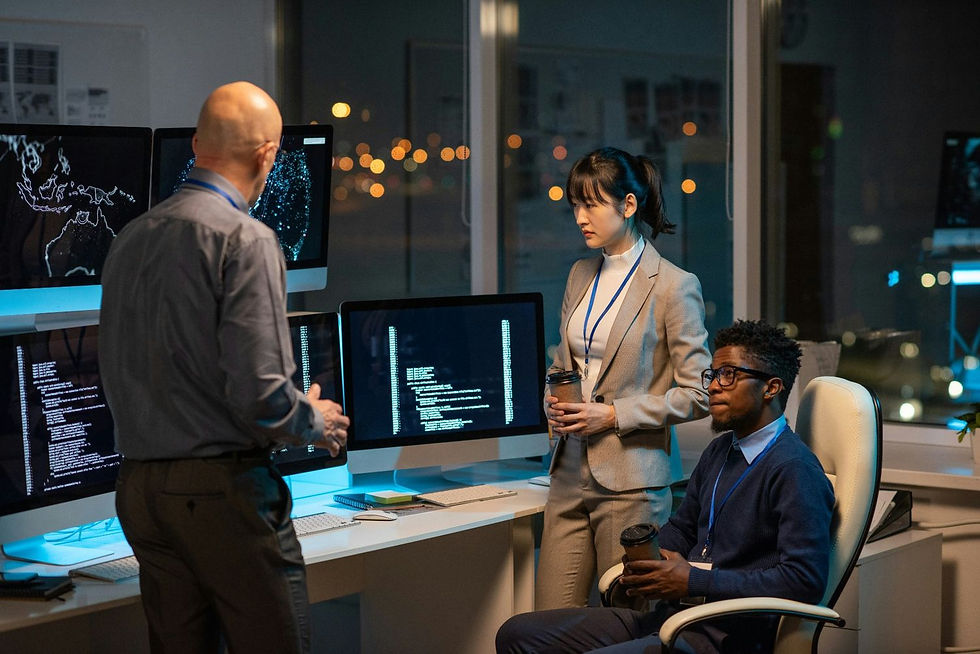 Three people in an office converse near screens displaying code. They hold coffee cups, creating a focused, professional atmosphere at night.
