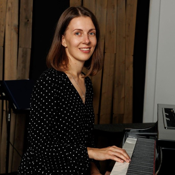Smiling woman in a black polka dot dress plays a piano indoors against a wood-paneled background.
