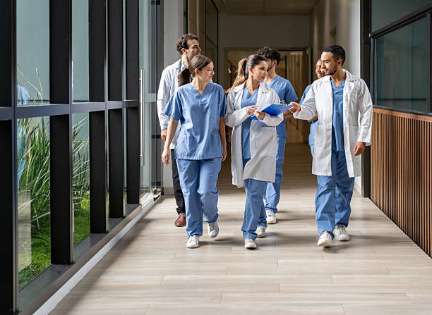 Group of healthcare professionals in blue scrubs and white coats walking down a hospital corridor, discussing and smiling. Bright setting.